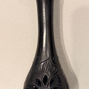May include: A tall, black vase with a slender neck and a flared top. The lower portion features intricate, carved floral and leaf patterns, with a central openwork flower design. The vase is approximately 30 cm tall, showcasing a blend of elegance and craftsmanship.