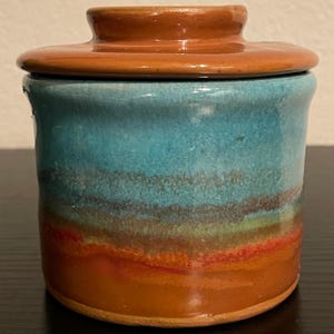 May include: A ceramic butter dish with a lid. The dish is cylindrical with a turquoise and brown glaze. The lid is a matching brown color. The glaze has a layered effect, with bands of color including red, green, and brown.