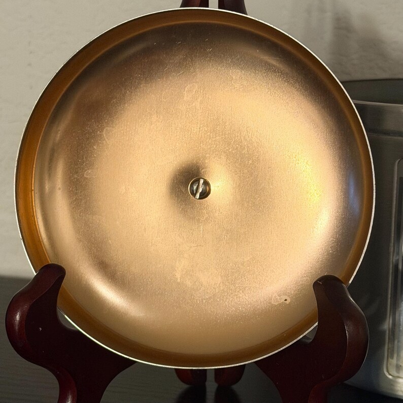 May include: A copper-coloured, round metal bell with a small screw in the centre. The bell is displayed on a dark wooden stand. The bell has a shiny, reflective surface and a slightly raised rim.