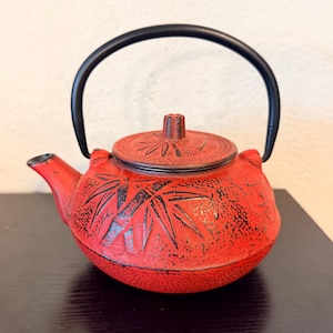 May include: A round, red cast iron teapot featuring a textured surface with a raised black bamboo leaf pattern. The teapot includes a matching red lid with a small knob and a black metal handle that arches over the top of the vessel.