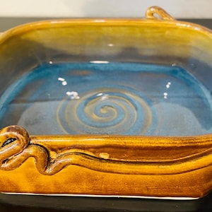 May include: A square ceramic baking dish with a blue interior and a spiral design. The exterior is a warm brown with curved handles. The dish is approximately 10 inches wide and 3 inches deep.