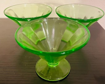 May include: Three vintage green glass dessert dishes. Each dish has a round bowl with vertical ridges and a pedestal base. The glass has a bright, translucent green color. The dishes are arranged on a dark surface.