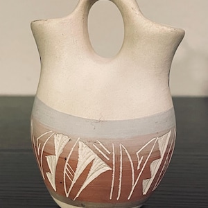 Może przedstawiać: Mały, kremowy ceramiczny wazon z charakterystycznym uchwytem i dekoracyjnym wzorem. Wazon posiada pas brązowo-szary z białymi geometrycznymi wzorami. Wazon to unikalny element ceramiki.