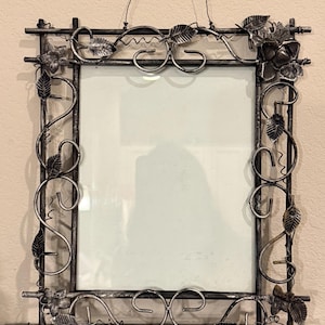 May include: A rectangular picture frame with a metallic silver and black finish. The frame features decorative metal scrollwork, vine-like accents, and sculpted leaves along the perimeter. A thin wire loop is attached to the top for hanging.