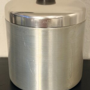 May include: A cylindrical, brushed metal container with a domed lid. The lid has a small, black, cylindrical handle. The container is silver and appears to be made of stainless steel. The container is sitting on a black surface.
