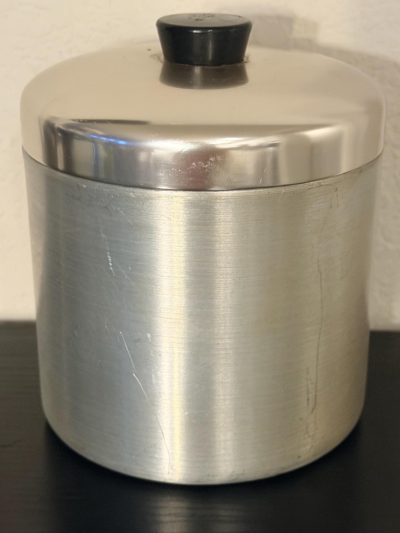 May include: A cylindrical, brushed metal container with a domed lid. The lid has a small, black, cylindrical handle. The container is silver and appears to be made of stainless steel. The container is sitting on a black surface.