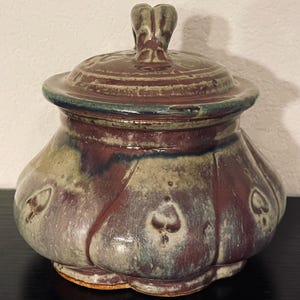 May include: A handmade ceramic jar with a lid. The jar is a mix of colors, including brown, beige, and burgundy, with a green rim. The lid has a handle and the jar has a heart-shaped design. The jar is a decorative item.