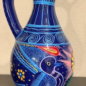 May include: A dark blue ceramic pitcher featuring hand-painted designs of a bird, a large pink flower, and yellow floral accents. The neck is decorated with light blue vertical lines and spiral patterns, while the handle is curved and solid blue.