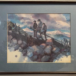 May include: Framed watercolor painting depicting a group of workers carrying beams across a rocky terrain. The artwork features a mountain range in the background and a cloudy sky. The painting is framed in a dark brown wooden frame with a blue mat.