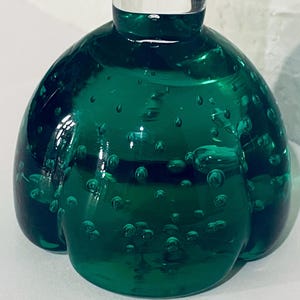 May include: A decorative, emerald green glass bottle with a clear glass stopper. The bottle's body is round with a bubbly texture, and the glass has a glossy finish. The design is reminiscent of a flower.
