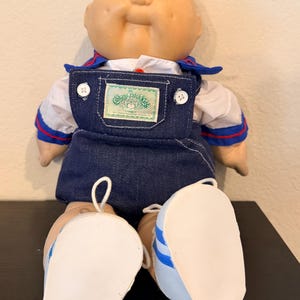 May include: A bald Cabbage Patch Kids doll with blue eyes and dimpled cheeks. The doll wears a white collared shirt with blue and red trim under dark blue denim overalls featuring a Cabbage Patch Kids patch and white shoes with blue stripes.