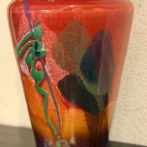 May include: A ceramic vase with a flared rim and a tapered body. The surface features a vibrant gradient of red, orange, and deep purple, accented by a textured green abstract line design and dark, mottled patches that create a layered visual effect.