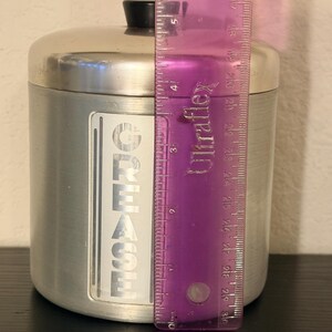May include: A silver metal container with a black lid. The container has a label that reads "GREASE" in vertical, bold, white letters. The container is approximately 12.7 cm tall.