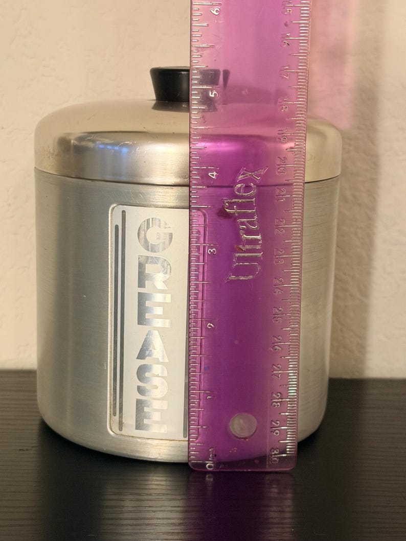 May include: A silver metal container with a black lid. The container has a label that reads "GREASE" in vertical, bold, white letters. The container is approximately 12.7 cm tall.