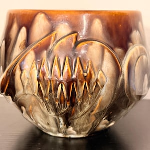 May include: A ceramic planter with a glossy, glazed finish. The planter features a rich brown color that transitions to lighter tones, with a textured, embossed design. The design includes stylized leaf and floral patterns.