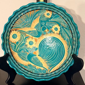 May include: A decorative, hand-painted ceramic plate with a teal and tan color scheme. The plate features a scalloped edge and a detailed design of birds, flowers, and foliage. The plate is displayed on a black stand.