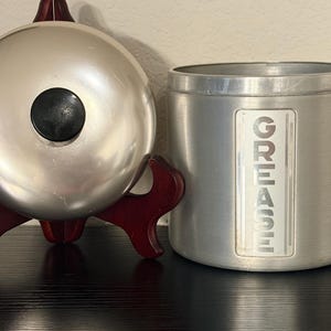 May include: A silver metal kitchen bell with a black centre button sits on a dark wooden stand. Beside it is a cylindrical metal container with the word "GREASE" printed vertically on the side.