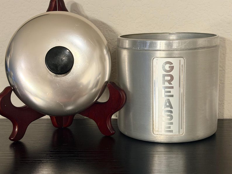 May include: A silver metal kitchen bell with a black centre button sits on a dark wooden stand. Beside it is a cylindrical metal container with the word "GREASE" printed vertically on the side.