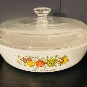 May include: White casserole dish with a clear glass lid and handle. The dish is decorated with colorful illustrations of vegetables and the text "Le Persil". A clear glass knob sits atop the lid. A vintage kitchenware item.