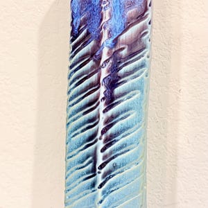 May include: A tall, slender ceramic vase with a V-shaped top opening. The surface features a textured, ribbed pattern with a glossy glaze that transitions from a light, pale blue at the base to a deep, vibrant violet and dark blue at the top.