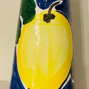 May include: A tall, ceramic vase with a hand-painted design. The vase features a large yellow lemon with green leaves against a dark blue background. The vase is approximately 12 inches tall.