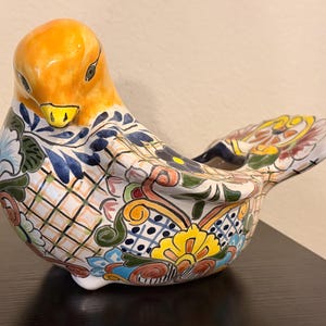 May include: A ceramic bird figurine featuring a bright yellow head and a body decorated with intricate hand-painted floral and geometric patterns in blue, green, red, and yellow. The bird has a glossy finish and a wide, flat tail section.