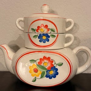 Op de afbeelding: Een stapelbare keramische theeset bestaande uit een theepot, een melkkan en een suikerpot met deksel. De witte stukken zijn voorzien van handgeschilderde bloemmotieven in rood, blauw en geel, omlijst door rode lijnen en groene bladeren.