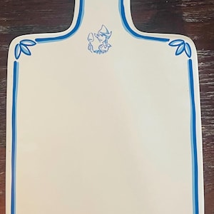May include: White rectangular cutting board with a blue border and handle. The board features a blue illustration of a person holding a wine glass and a cheese wedge design. Decorative leaf accents are in each corner.