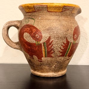 May include: A textured brown clay pitcher featuring a wide handle and a flared rim with a yellow painted border. The body of the vessel is decorated with two stylized red bird figures, each accented with green and yellow vertical stripes on their wings.