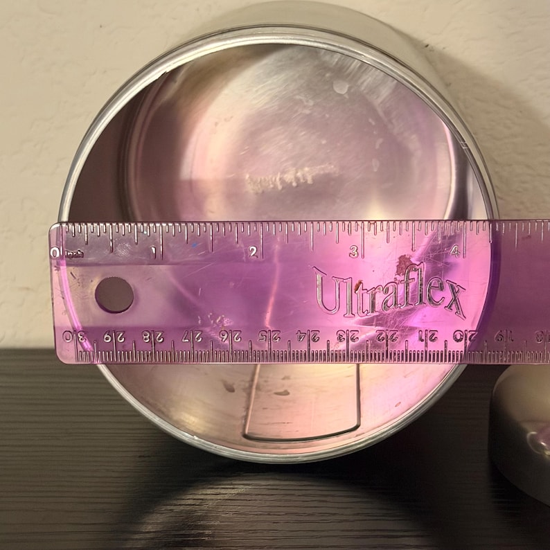 May include: A silver metal container with a ruler measuring its diameter. The ruler, marked in centimetres, shows the container's diameter is approximately 7.6 cm. The word "Ultraflex" is printed on the ruler.