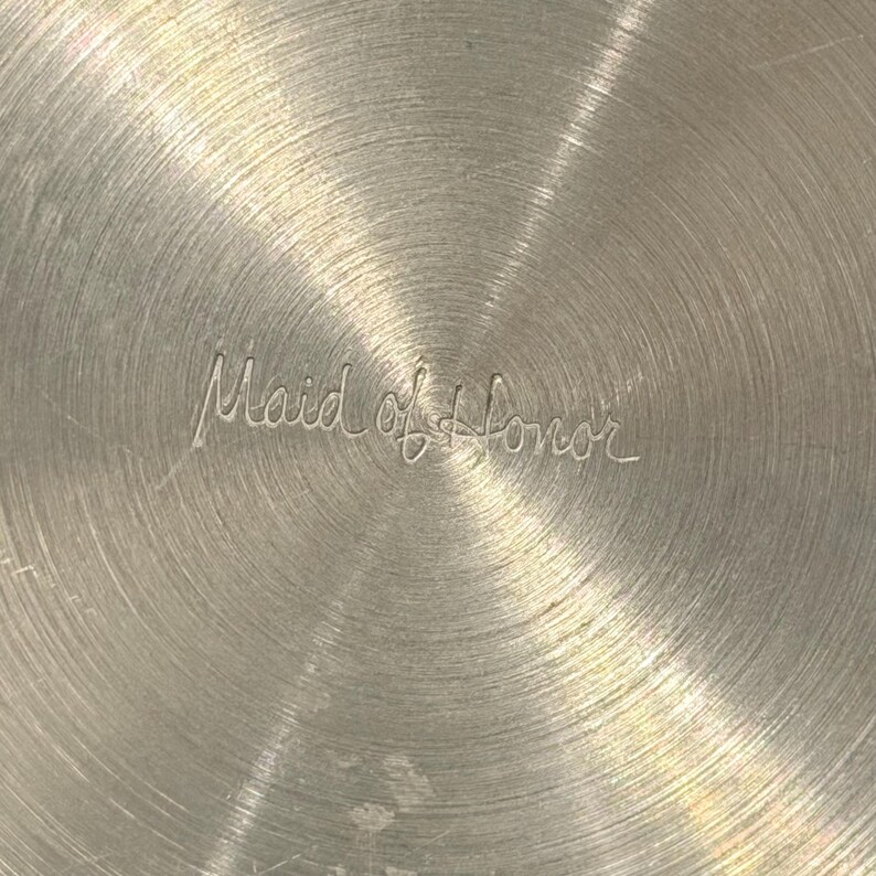 May include: Close-up of a silver metal surface with a circular brushed pattern. The engraved text "Maid of Honor" is centered, suggesting a decorative item for a wedding or special occasion.