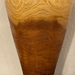 May include: A wooden vase with a flared top and a rounded base. The vase has a natural wood grain pattern with a gradient of light to dark brown tones. The vase is a decorative item, likely for displaying flowers.