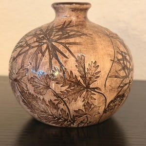 May include: A round ceramic vase with a tan, textured finish featuring dark brown botanical illustrations of leaves and stems. The vase has a narrow neck and a smooth, glossy glaze over the detailed plant patterns etched into the surface.