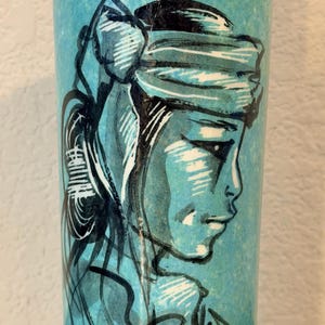 May include: A cylindrical turquoise vase featuring a black and white ink-style illustration of a person in profile. The figure wears a headband and has long hair tied back. The vase has a mottled, textured finish with a smooth, glossy appearance.