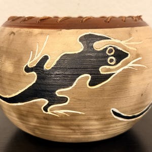 May include: A round, tan-colored ceramic bowl featuring a dark brown rim with visible stitching. The exterior surface is decorated with black lizard silhouettes outlined in white, set against a textured, light brown, horizontally-lined background.