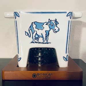 May include: White and blue ceramic fondue pot with a cow illustration. The pot has a square shape with handles and sits on a wooden base. The base has the text "BOSKA® HOLLAND". The pot has a metal insert with star cutouts.