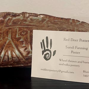 May include: A brown, textured ceramic piece with etched geometric patterns sits behind a white business card. The card features a hand logo, the text Red Deer Pottery, Sandi Fanning Potter, wheel thrown and hand built stoneware and raku pottery.
