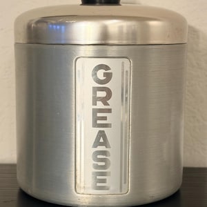 May include: A cylindrical, brushed metal container with a lid and a black knob. The word "GREASE" is vertically printed in white on a rectangular label on the front. The container is designed for kitchen use.