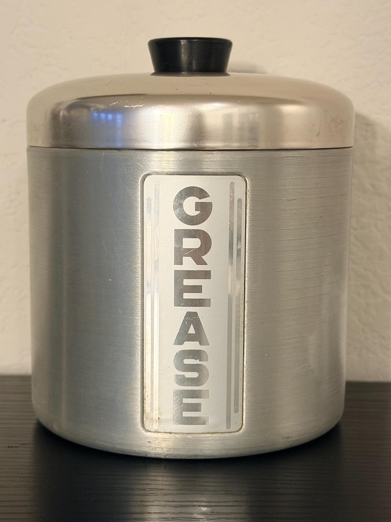May include: A cylindrical, brushed metal container with a lid and a black knob. The word "GREASE" is vertically printed in white on a rectangular label on the front. The container is designed for kitchen use.