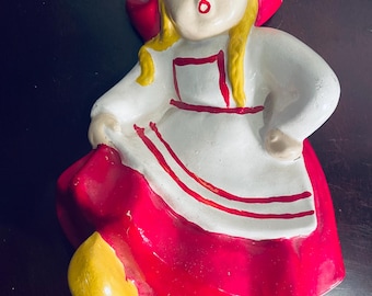 May include: A ceramic figurine of a girl wearing a red dress and a red and white hooded cloak. She has blonde hair and is wearing yellow shoes.