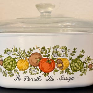 May include: A white rectangular casserole dish with a clear glass lid and side handles. The front features a colorful botanical illustration of vegetables and herbs, including a tomato and mushrooms, with the text Le Persil La Sauge below.