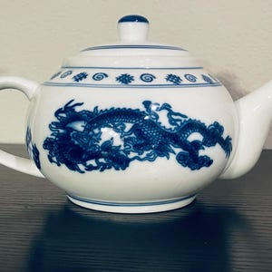 May include: White ceramic teapot with a blue dragon design. The teapot has a rounded body, a curved handle, and a spout. The lid is topped with a small blue knob. The dragon is depicted in a detailed, stylized manner, wrapping around the body.
