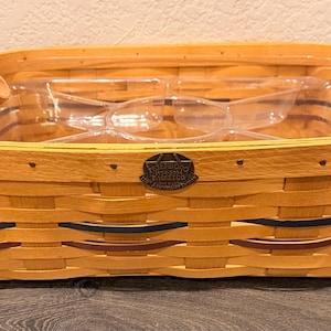 Może przedstawiać: Prostokątny pleciony kosz z brązowymi skórzanymi uchwytami i akcentami. Kosz ma jasnobrązowy kolor z ciemnoniebieskimi i brązowymi akcentami. Wewnątrz kosza znajduje się przezroczysta plastikowa wkładka. Metalowy emblemat znajduje się z przodu.