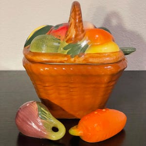 May include: A ceramic basket-shaped container with a handle, filled with colorful fruit-shaped lid. Includes a carrot and eggplant-shaped salt and pepper shakers. The basket is a warm orange color, and the fruit on the lid includes green, red, and yellow elements.