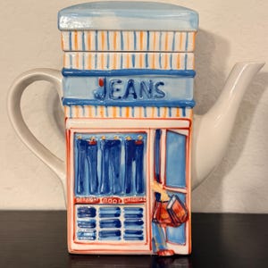 May include: A white ceramic teapot with a unique design resembling a jeans store. The teapot features the word "JEANS" in blue lettering, with blue jeans displayed in the windows. The design includes orange and blue stripes.