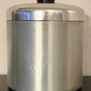 May include: A brushed aluminium canister with a rounded lid and a black knob. The cylindrical container has a vertical brushed texture and a few scratches. The lid is also brushed aluminium.