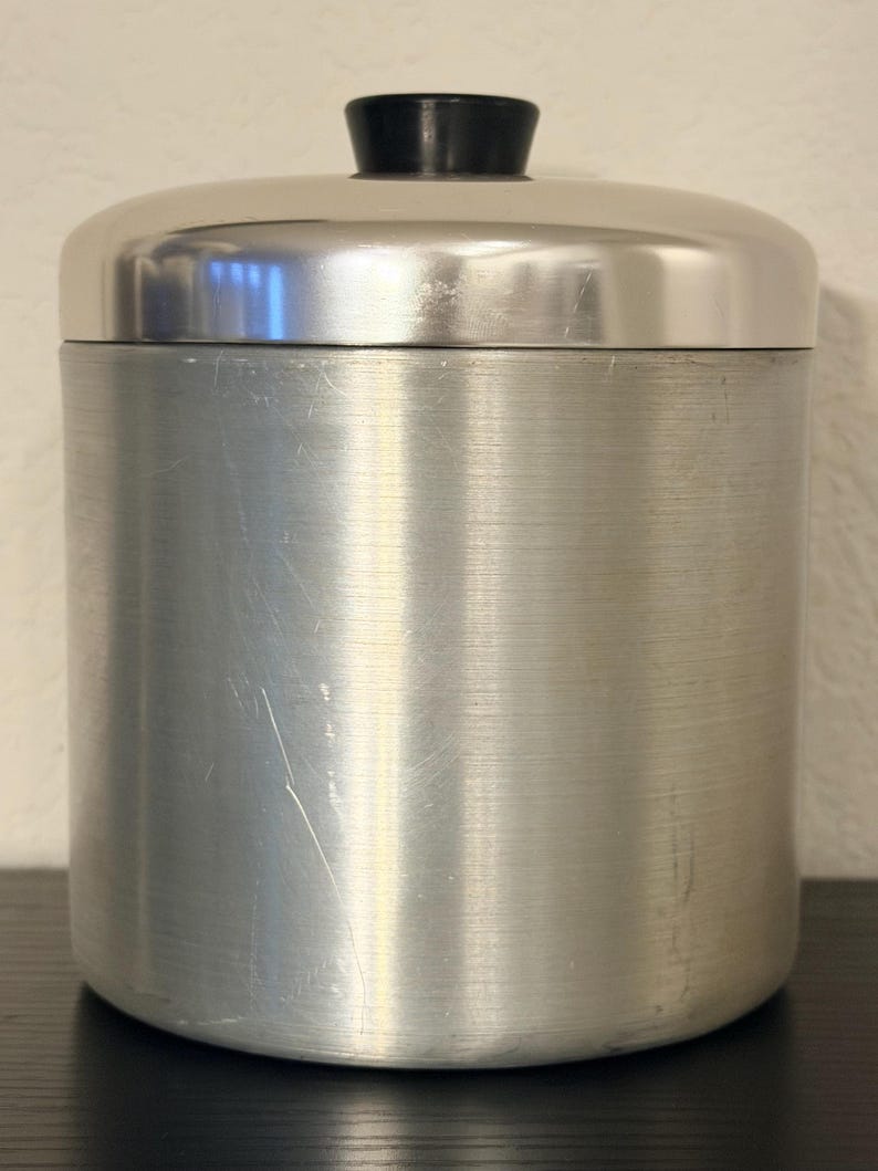May include: A brushed aluminium canister with a rounded lid and a black knob. The cylindrical container has a vertical brushed texture and a few scratches. The lid is also brushed aluminium.