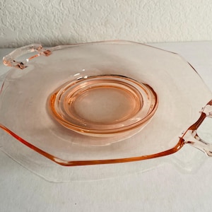 Puede incluir: Un plato de servir de vidrio rosa con dos asas. El plato tiene un borde festoneado y un centro elevado.