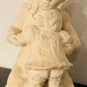 Op de afbeelding: Een crèmekleurige keramische sculptuur van een moeder die een kind omhelst. De figuren zijn afgebeeld met gesloten ogen en zachte kenmerken. Het kind draagt een jurk met ruches en kniehoge sokken. De sculptuur heeft een matte afwerking.