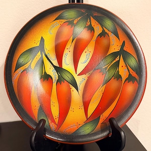 May include: A round decorative plate featuring a vibrant hand-painted design of multiple red chili peppers on green stems with leaves. The background transitions from a bright yellow center to a deep orange and black border around the edge.